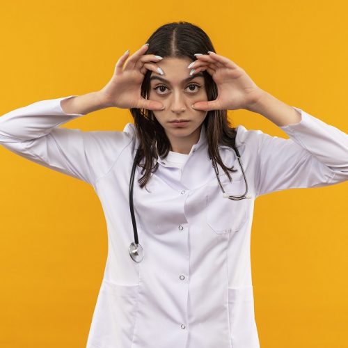 young woman doctor in white coat with stethoscope around her neck opening eyes with fingers feeling morning fatigue standing over orange background